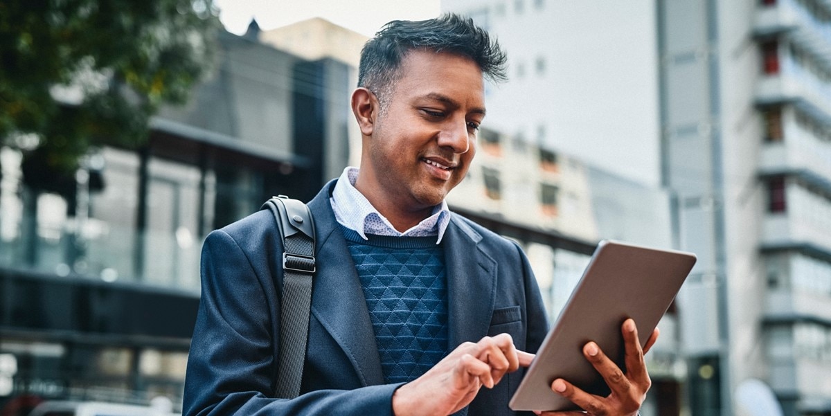 man standing outside building with bag on shoulder, looking at tablet in hand